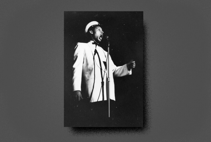 A black-and-white photo of Ernie Andrews in performance shows him standing at a microphone with his left arm raised. He is wearing a white jacket, shirt and hat, with a black bow tie.