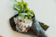 A hand roll wrapped in nori, filled with rice, dry-aged beef, melted cheese with visible char marks, and topped with a bundle of fresh arugula, photographed close-up against a white background.
