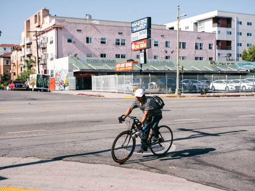 A cyclist rides on a street where there is no bike lane. He is wearing a cap but no helmet and he has a backpack. Businesses and apartment buildings can be seen in the background.