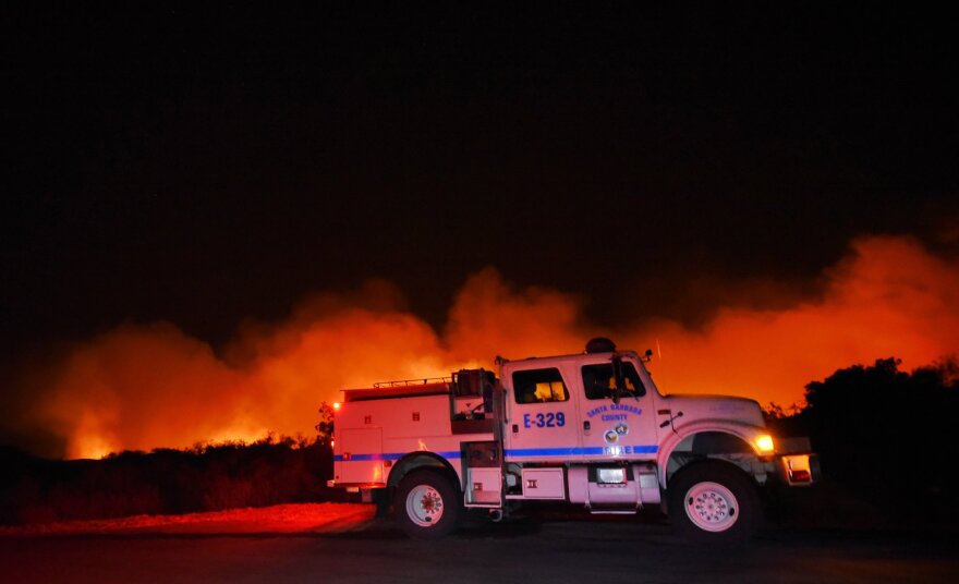 A fire is shown burning land at night. In the foreground, a white and blue Santa Barbara County fire truck is parked. In the background, bright red, orange and yellow flames can be seen against a black sky.