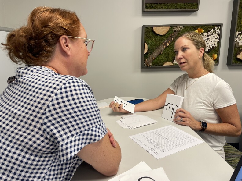 Two women sit across from each other at a table. One is holding up a flashcard with the letters -mp. A sheet of paper with many lines and boxes lies between them, along with a pile of flashcards.