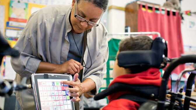 A woman in glasses helps a young boy in a wheel chair with navigating a computer screen.