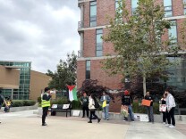 A small group of students stands outside a brick building. A Palestine flag is raised neared them. One student in a yellow vest holds a bullhorn.