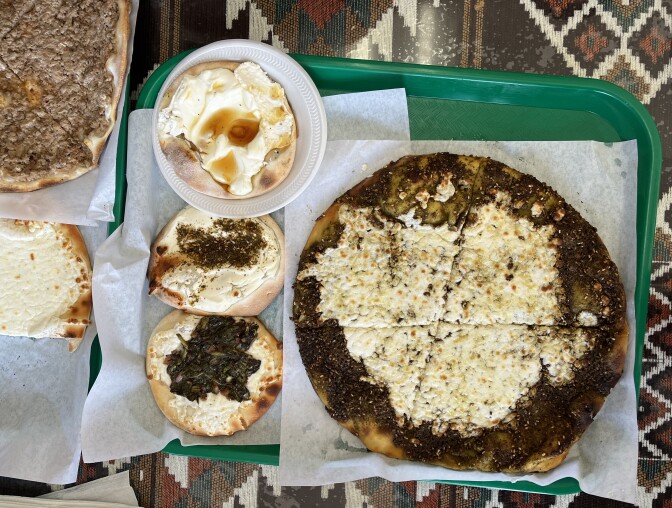 An overhead image of various flatbreads situated next to each other, all with different types of cheese and seasoned toppings. The flatbreads sit on a green tray placed upon a clear glass tabletop.
