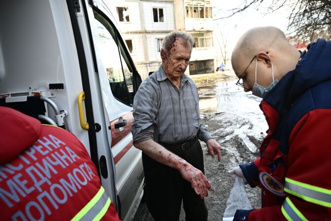An older man with thinning gray hair has blood on his head and his arm. First aid workers attend to him in front of a truck, with puddles and another building in the background.
