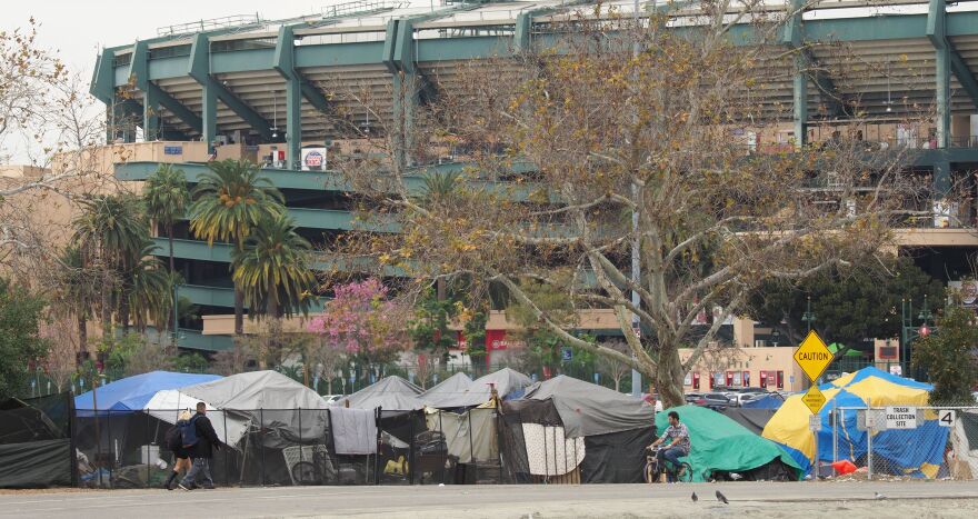 Tents and tarps are situated side-by-side in a homeless encampment on a sidewalk outside Angel Stadium, which is seen in the background. A bicyclist rides by the encampment in the foreground.
