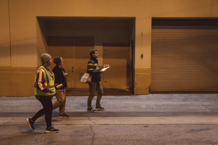 A white woman with a safety vest, black pants, and a gray beanie walks next to a person with a black jacket and sweats and a white man with a puffer jacket, jeans and a satchel. They walk by a brown and white wall. It's night time.