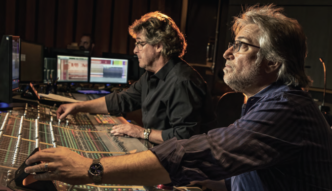 Two male presenting people in front of a console that looks like an audio mixing board.