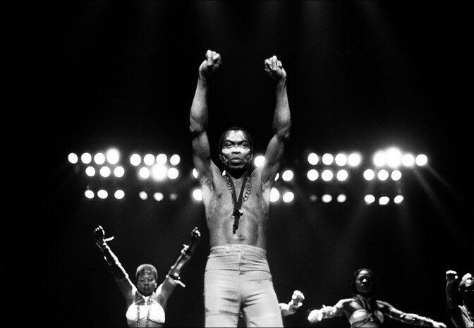 Black and white photo of a shirtless man, wearing light colored pants raising both his fists in the air as he stands on a stage. A row of three women are standing behind him, also with their hands in the air.