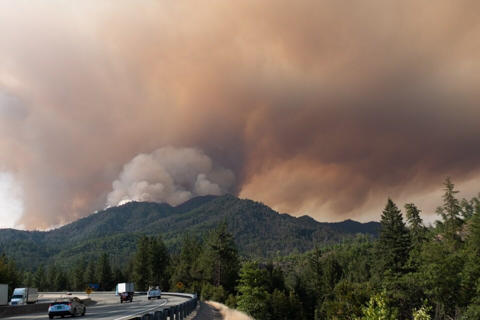 A massive plume of smoke rises from green hills as cars drive on a windy road in the foreground