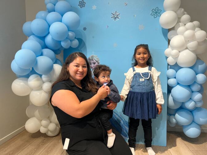 A young female wearing a blue and white dress stands in front of a blue and white balloon and panel backdrop beside a woman wearing all black holding a young child. 