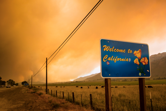 An orange/gray smokey sky with a sign in the foreground that reads "Welcome to California"