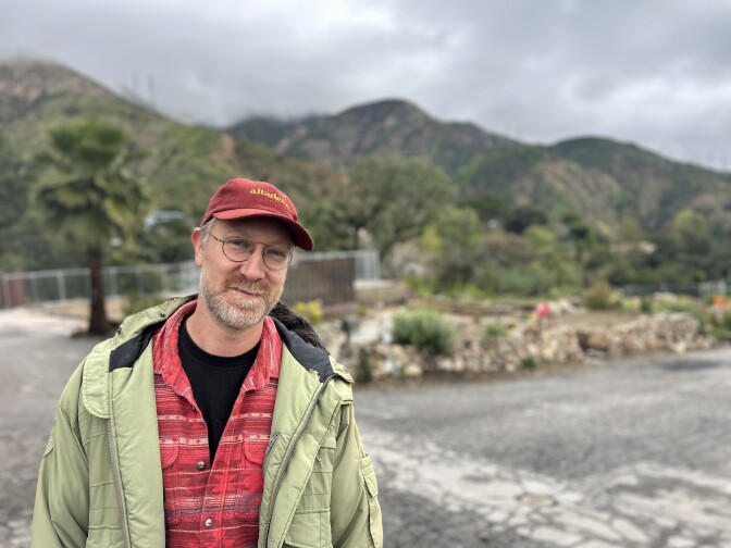 A middle aged man with light skin and a short mustache and beard wears round glasses, a red baseball cap and light green jacket.