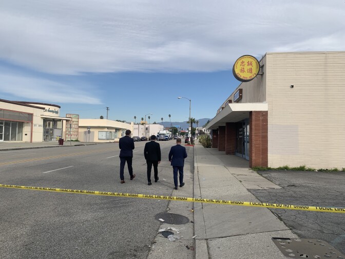Three people in dark suits walk down the street next to buildings behind the yellow do not cross tape during the day.