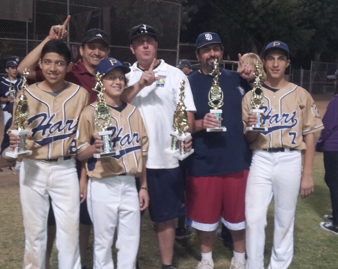 Three boys in beige baseball jerseys that read "Hart" pose with three men. All hold trophies. Two of the men are the left are signaling "No.1" with their fingers.