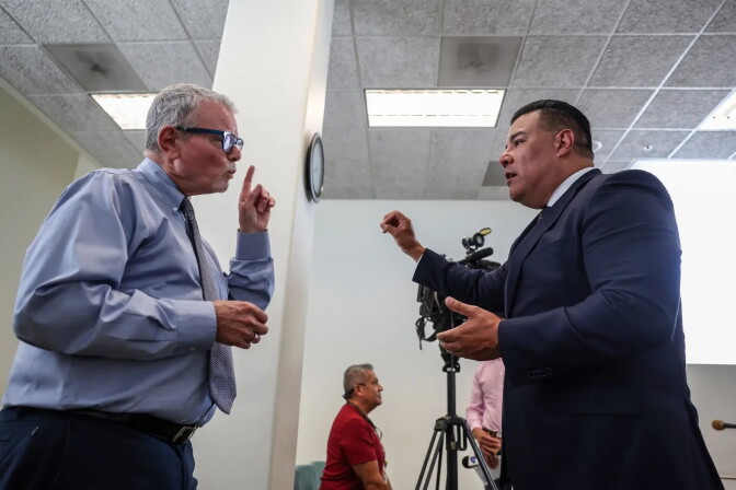 Two men in business attire gesture while talking closely, with a camera crew and others in the background.
 