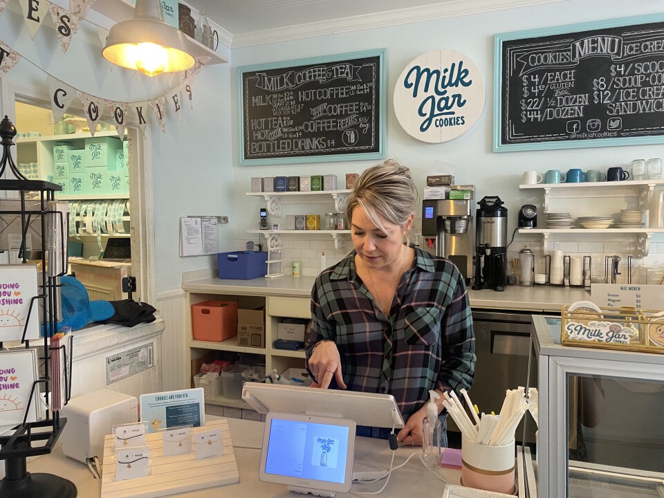 A grey-haired light-skinned woman in plaid takes cookie and coffee orders at the front desk of 'Milk Jar Cookies.'