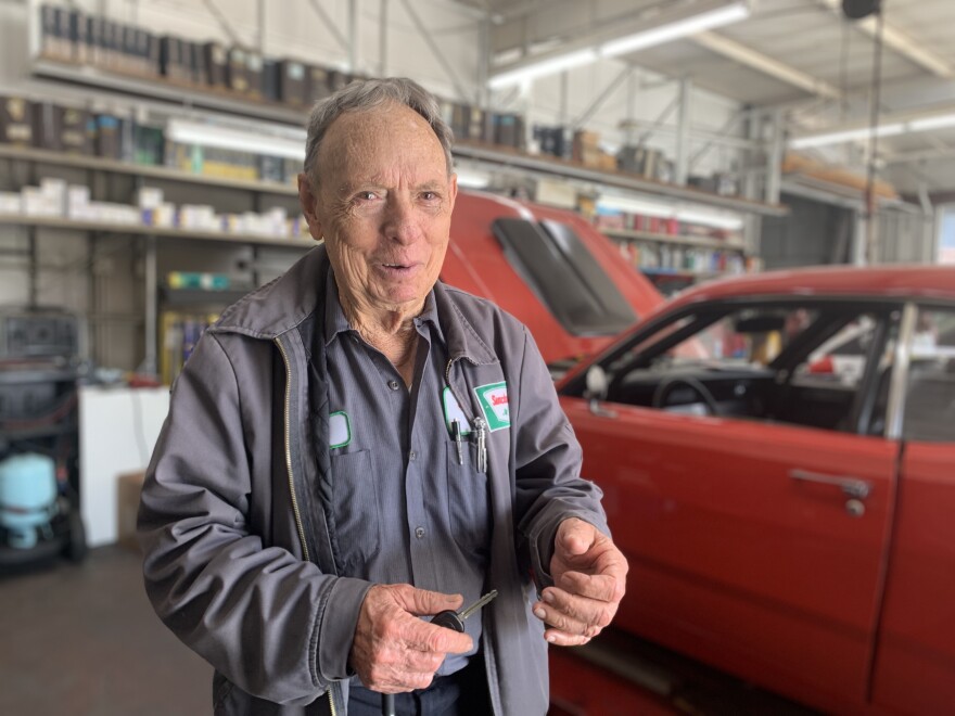 An older man with light skin tone in an auto repair shop uniform stands in a body shop with a red car behind him.