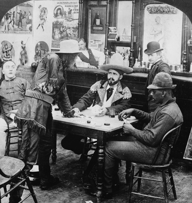 A man in a fringed leather jacket and cowboy hat holds a revolver at his waist as he argues with a seated fellow gambler.