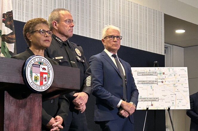 A woman with very short curly hair is standing next to a podium with the City of Los Angeles emblem on it. Next to her, a man in a law enforcement uniform and another man in a blue suit stand solemnly next to a map of the city with three marked locations on it. 