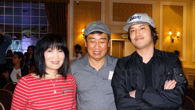 An Asian woman with light skin tone and short black hair with bangs wearing a red and white striped shirt stands next to an Asian man with medium-light skin tone wearing a gray shirt and hat and glasses standing next to an Asian young man with a gray Lakers baseball cap and black jacket. 