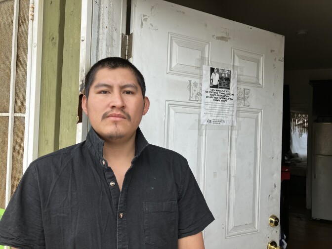 A male-presenting person with medium to light skin tone, short dark hair and a mustache, wearing a black shirt, stands before a white-painted open doorway that has a flyer posted on it.