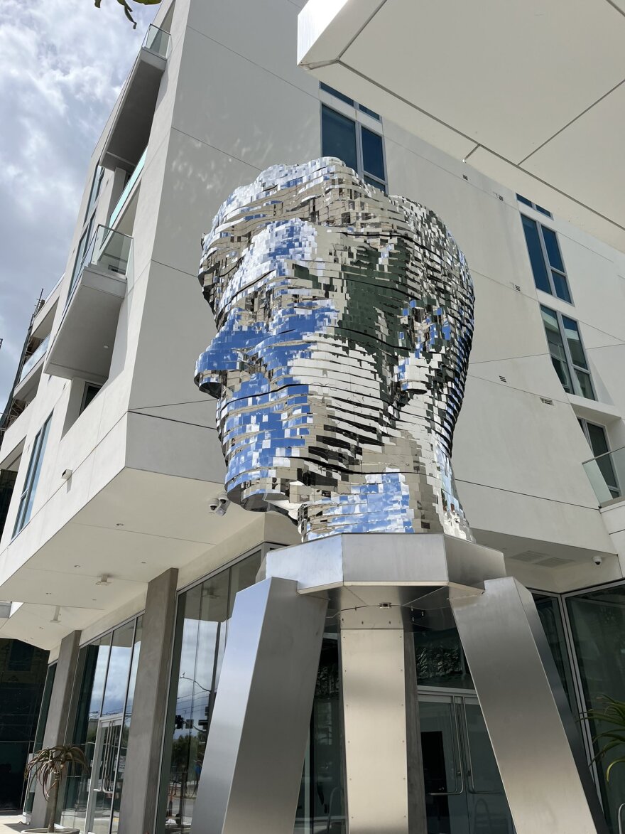 An art installation of a giant, shiny silver metal head of David Lynch, a renowned film director. The head is displayed on a slightly less shiny, but still silver pedestal between two large white buildings with balconies and windows.