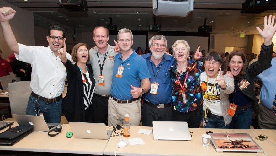 A group of nine people stand at a long table celebrating with hands in their air. Many wear orange ID badges around their necks.