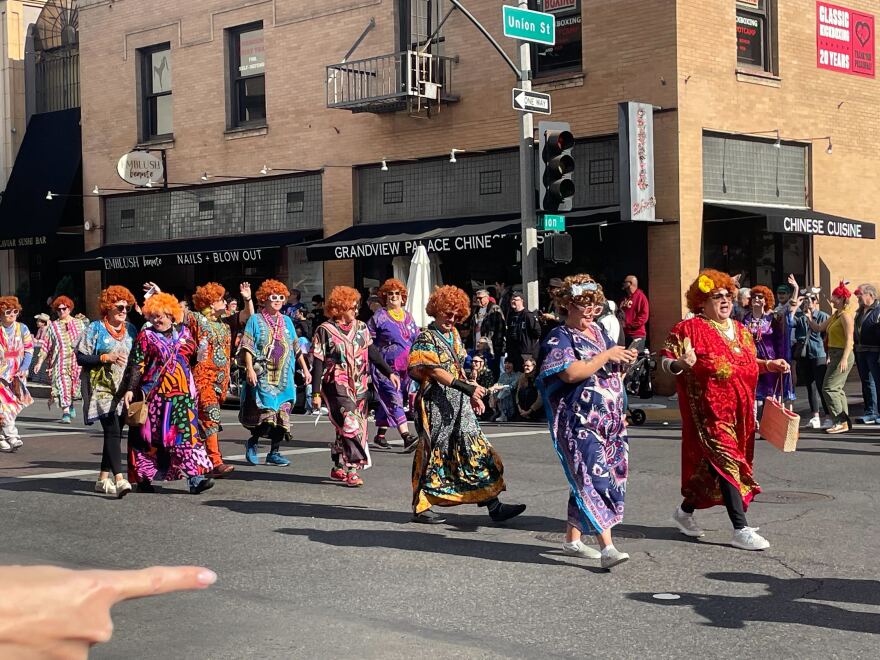 A group of women in orange curly wigs and colorful print dresses, in the style of the character Helen Roper from "Three's Company," walk down the street in a parade.