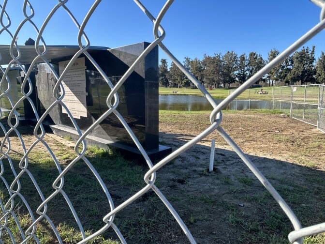 Behind a chain-link fence, a long black wall with a tarp over it sits on a packed mud and grass piece of land with a small lake in the background. 