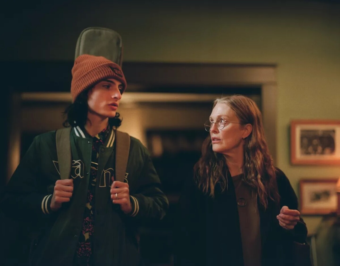 Finn Wolfhard and Julianne Moore stand in the entry way of suburban house. The room is lit with warm yellow lights giving the walls a light green hue. Picture frames can be seen hanging on the wall behind Moore to her left and the living room can be seen behind her and Wolfhard out of focus. Wolfhard stands to Moore's right. He wears a green bomber jacket as he holds the straps of his backpack tight on his chest. The stem of a guitar can be seen poking out from behind him. Wolfhard also wears an orange beanie, his black wavy hair just meeting his shoulders. Moore wears a brown t-shirt covered by an overside black cardigan. She holds a handbag hanging from her left arm at the elbow. Moore also wears round wire glasses with her hair loosely falling past her shoulders. 