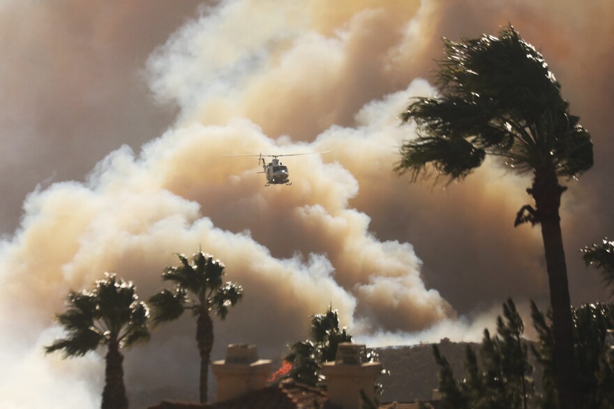 Palm trees are bent from the wind as a helicopter flies above homes threatened by the wind-driven Palisades Fire in Pacific Palisades.