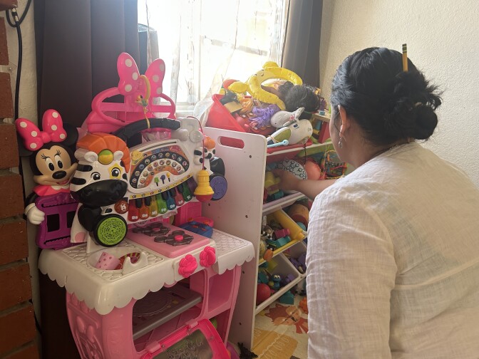 A woman with medium-light skin tone tidies up a shelf teeming with toys. Next to the shelf, there is a pink play kitchen, along with a toy piano in the shape of a zebra. 