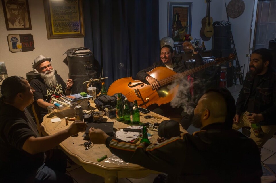A group of men sit around a table and laugh. One of them holds a large stringed instrument.