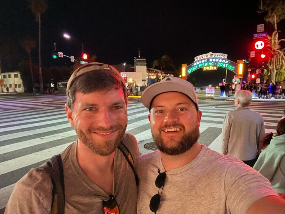Two men, both wearing hats, pose in front of a beach pier at night