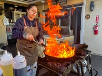A woman in a brown Lomo Fuego apron stirs a wok over a powerful outdoor burner, producing dramatic flames that leap several feet into the air in a backyard restaurant's  patio area. 