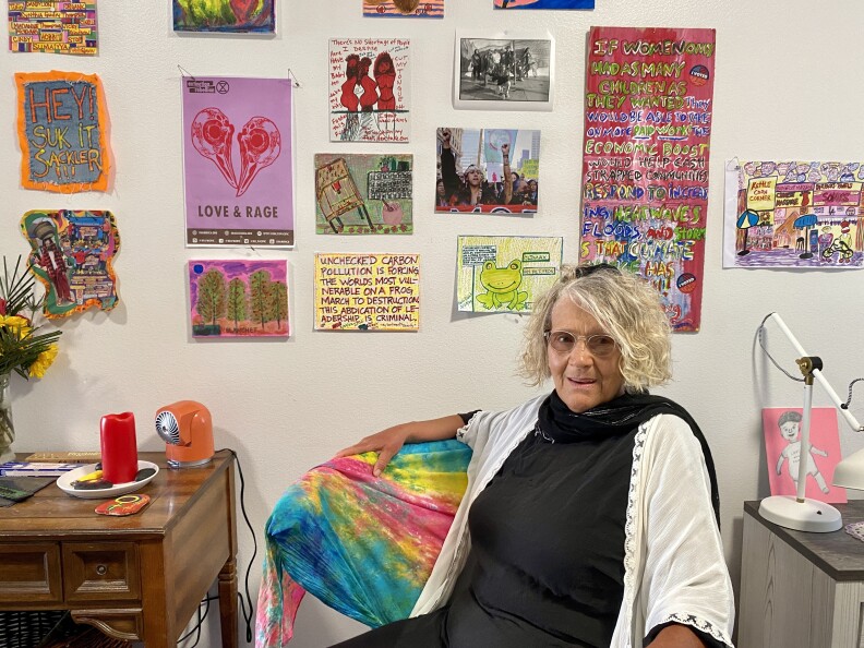 Evans is sitting in a chair inside her apartment. There is a wall of art behind her of dozens of art she's collected or made over the years.