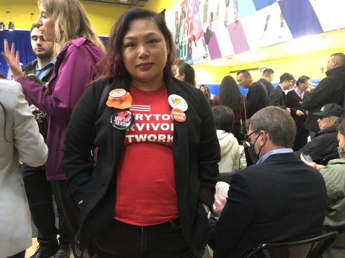 An Asian woman wears a red t-shirt with partially obscured white lettering and a light black jacket with four large buttons pinned to it