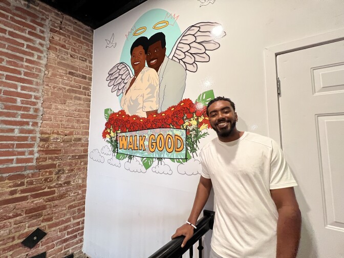 A brown skinned man stands in front of the mural of his grandparents. 