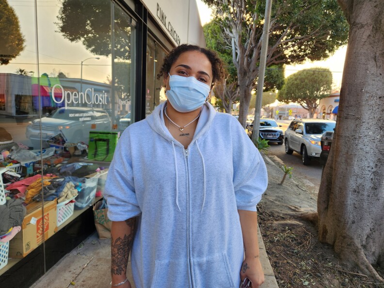 A medium skinned young woman wearing a blue mask and a blue zip up hoodie, wearing a silver necklace, stands on a street next to a tree in front of a store window that says OpenClosit