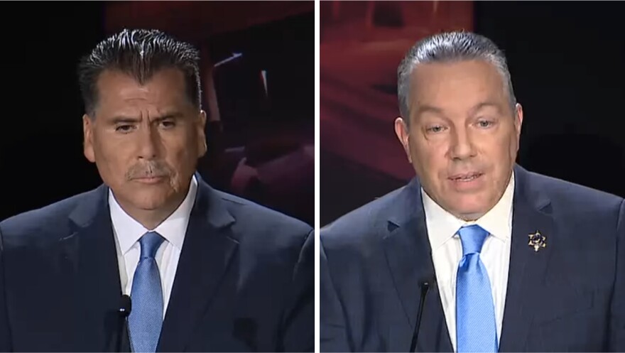 Robert Luna and Alex Villanueva stand at their respective debate podiums, both dressed in navy suits with light blue ties.