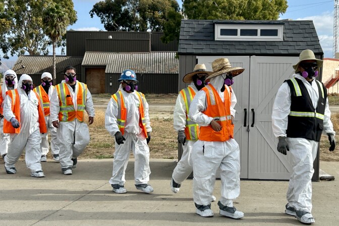 A line of workers wearing white protective suits, orange safety jackets and protective breathing masks walks across a concrete slab. 
