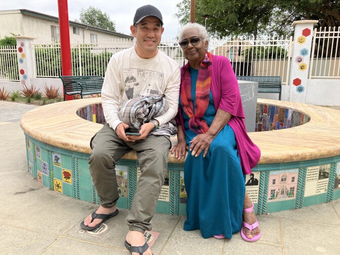 A man in a baseball cap, white graphic t-shirt, olive green pants and flip-flops, left, sits at a fountain with an elderly woman, right, who is wearing a long blue dress and fuschia jacket and scarf.