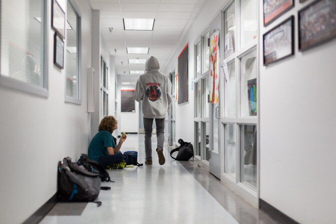 Two students in a bright school hallway: one sits on the floor against the wall eating a snack beside backpacks and a lunch bag, while another in a gray hooded sweatshirt walks away from the camera.