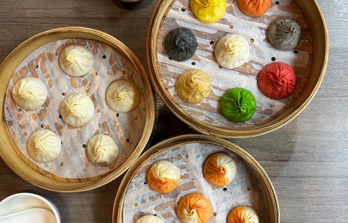 A photograph of three sets of dumplings in bamboo steamer baskets, each a different color, sitting on a wooden table. The bottom group of dumplings is orange and white. The center group is white. The upper right basket contains dumplings that are green, red, grey, back, yellow, orange and white. 