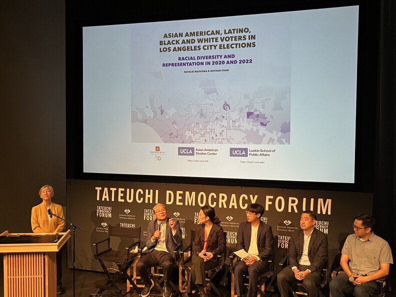 A photo of four men and one woman seated inside a hall with a map projected behind them entitled "Asian American, Latino, Black and White Voters in Los Angeles City Elections." A woman stands at the podium. 