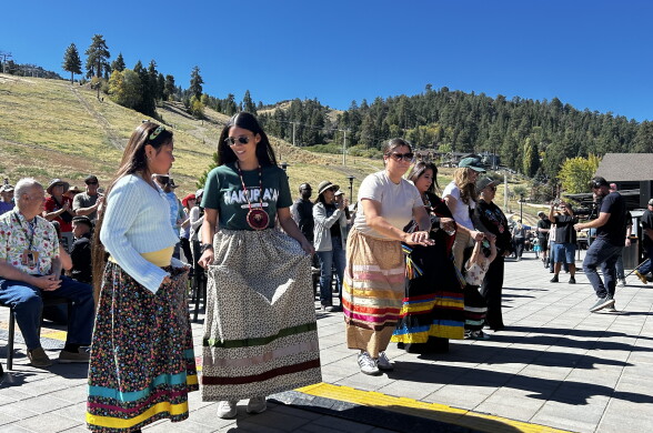 People in long patterned skirts hold hands in a line dance outdoors, with pine-covered hills in the background.