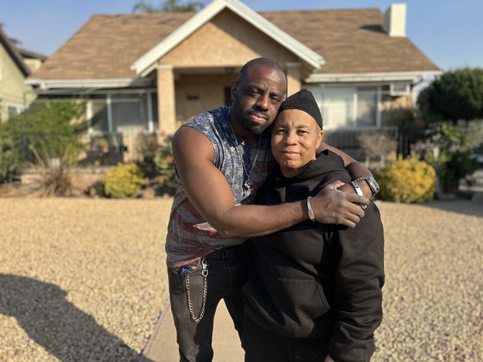 A younger man with dark skin tone wearing a sleeveless American flag shirt and black pants with a chain hugs a middle-aged woman with medium dark skin tone wearing a black sweatshirt and durag in front of a modest, tan single-family home. It's sunny and they both have slight smiles looking at the camera. 