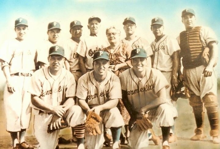 10 men all wearing baseball uniforms stand or kneel. One woman in a dress stands among them.