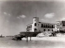 A black and white photo depicts a beachfront marine lab with a central tower and tiled roof. 